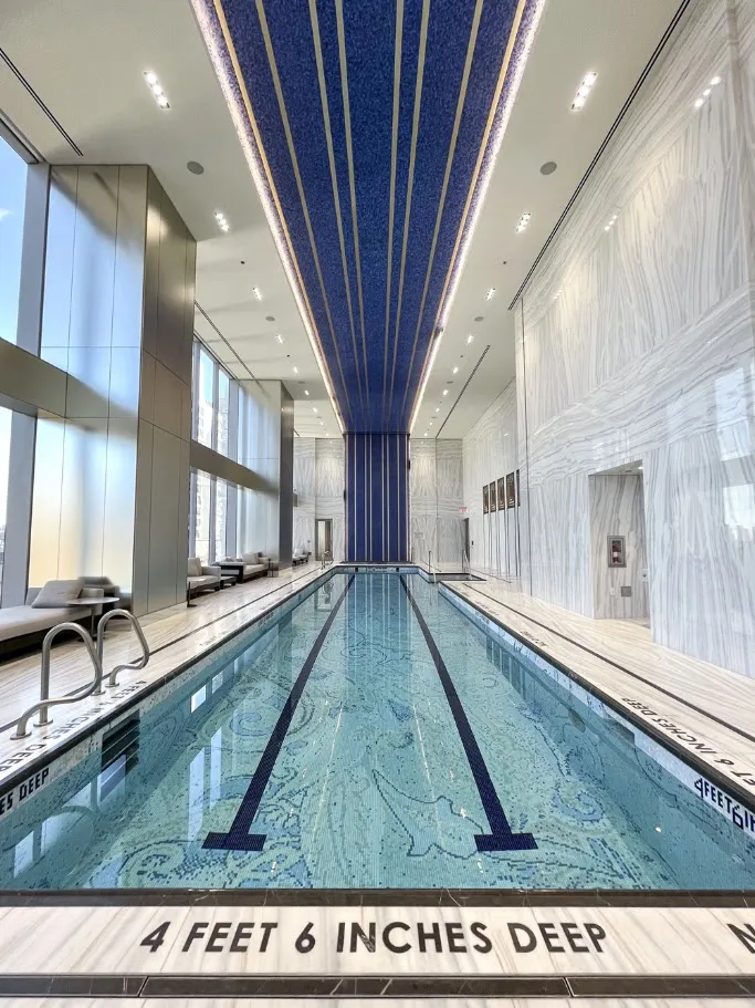 High-end indoor lap pool featuring intricate custom blue mosaic basin tiles, striking white linear-veined marble-look deck tiles, and floor-to-ceiling windows.