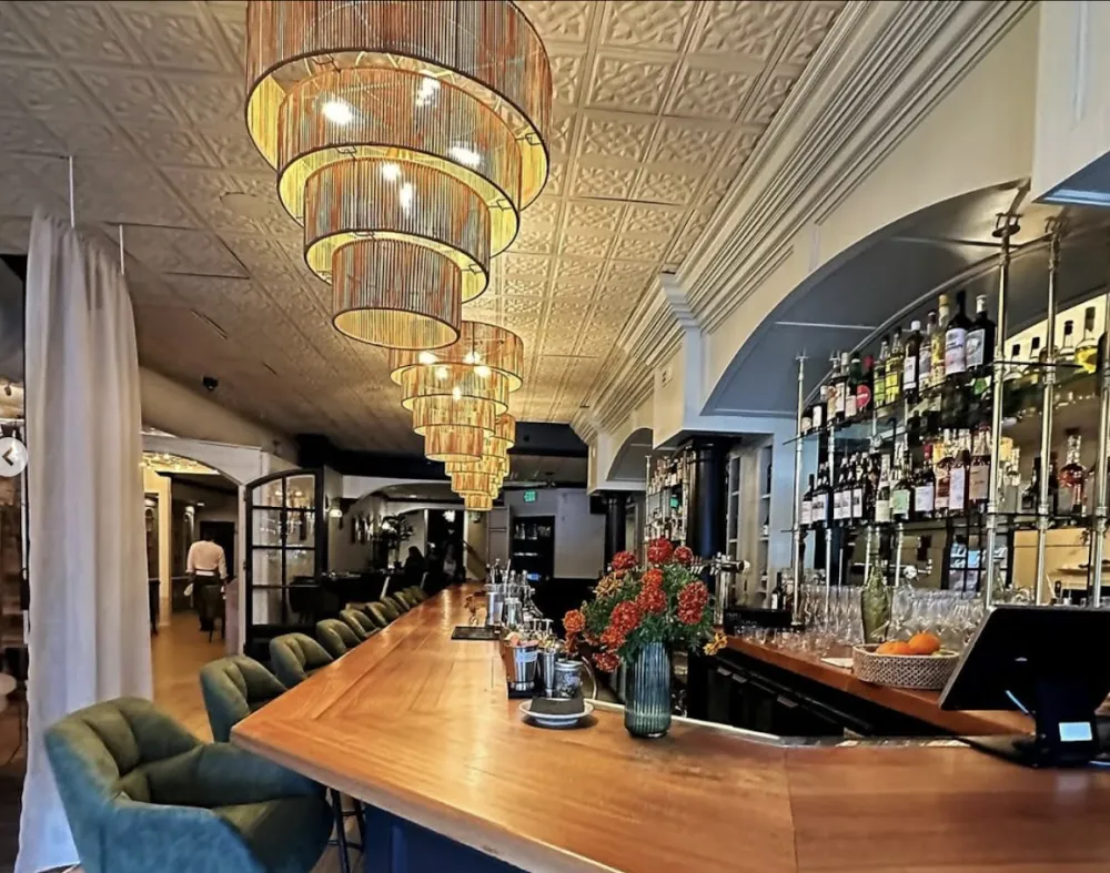 Upscale restaurant bar designed by Kerri-Lee Mayland featuring a wood counter, olive green velvet bar chairs, and embossed ceiling tiles.