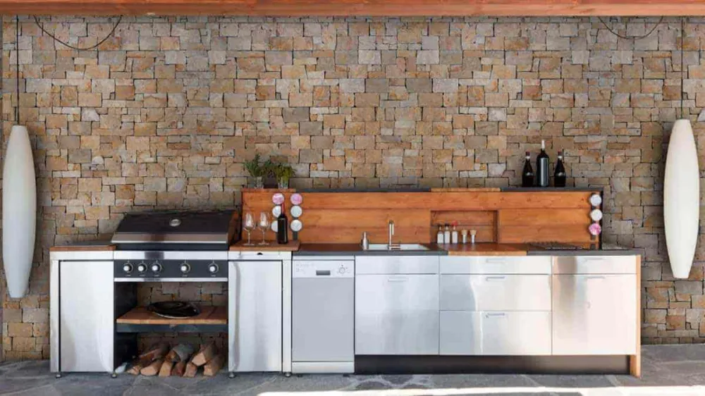 Outdoor kitchen with latte-toned stone cladding, stainless cabinetry, grill, wood countertop, and covered patio wall.