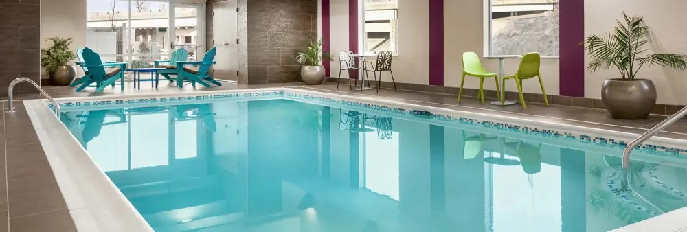Modern hotel indoor swimming pool featuring dark grey porcelain deck tile, white coping, blue glass mosaic waterline tile, and colorful lounge seating.